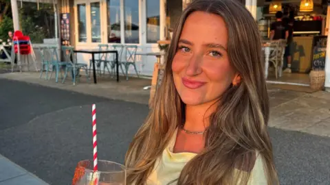 Molly Lambert A young woman with long brown hair smiles at the camera. It is a close-up of her, showing her head and shoulders. She wears a light yellow top and holds a cocktail.