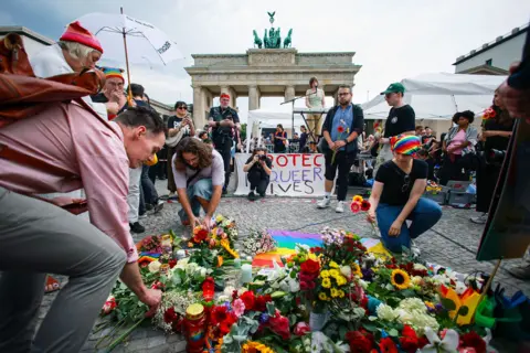 Clemens Bilan / Reuters People lay flowers in front of a banner saying 'protect queer lives', with the Brandenburg Gate in the background on a cloudy day in Berlin on Sunday. 