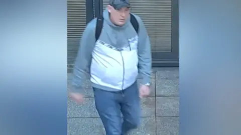 The image shows a man walking outdoors on a paved surface. He is wearing a light grey and white hooded jacket, blue jeans, black shoes with white accents, a dark backpack and a dark cap.