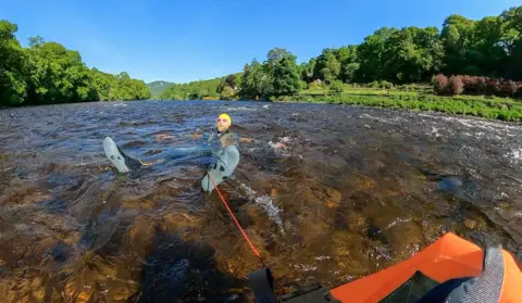 Calum Maclean Wild swimmer Calum floating in the River Tay between Dunkeld and Kinclaven on a sunny day in May this year. His head and feet are sticking out of the water. He's wearing water shoes and a bright yellow swim cap and dark goggles.
