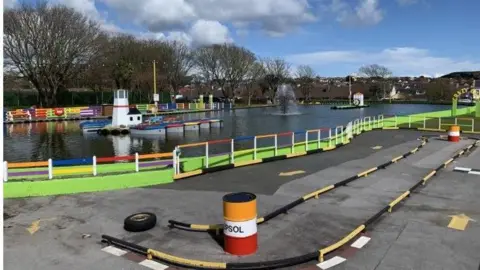 A wide shot of the Onchan Park lake. There are several small boats in the water. The water is surrounded by multi-coloured railing.