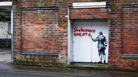 King's Lynn and West Norfolk Council A wider view of art painted on white double doors on a brick building. The art shows an image that looks like William Shakespeare from behind in black. He is holding a spray can and the words 'Shakespeare woz ere' have been written in red ink