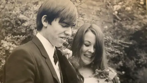 Family photo John wears a dark suit with a carnation buttonhole. Tina is wearing a white long-sleeved dress with a large floral buttonhole. They are smiling and looking down at their marriage certificate.