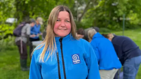 A woman with long blonde hair, wearing a blue jacket, looks at the camera