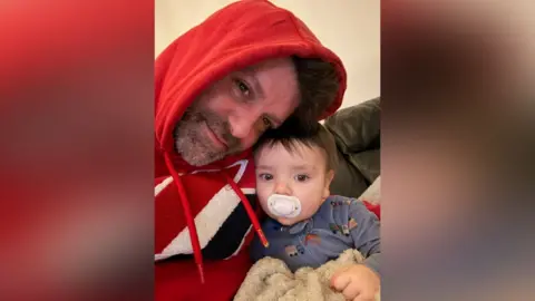 Family photograph of Gary Pidgley with his young son. Gary is wearing a red hoodie and is cuddling his son, who is holding a blanket and has a dummy in his mouth.