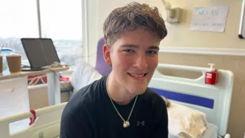 Hamish looks at the camera and smiles. He is wearing a black t-shirt and we can see his upper torso and head only, he is wearing a gold ring on a chain around his neck. In the background we can see his hospital room including the bed and table as well as a window beyond. 