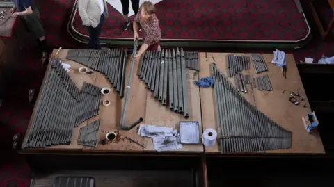 Around one hundred pipes which have been removed from the pipe organ lay on a large table in front of the organ. Volunteers dust and clean them with paint brushes and cleaning products. 