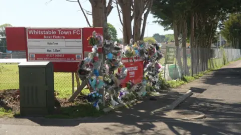 Floral tributes are attached to a fence outside Whitstable Town FC, beside the club’s red sign and entrance road. The fence runs alongside a row of trees and a pitch can be seen behind the fence.