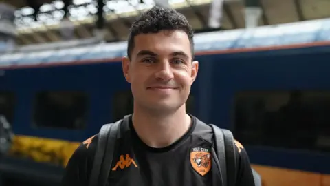 Jasmine Lowe/BBC John Egan who has short black hair and is wearing a black Hull City T-shirt stands on the platform at Hull station
