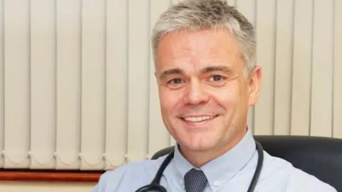 North East and North Cumbria ICB Dr Neil O'Brien is sitting in a black chair and smiling at the camera. He has short greying hair and is wearing a blue shirt and tie. There is a black stethoscope draped around his neck.