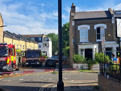 A house with a blown out window on a residential street, with fire engines parked along the road 
