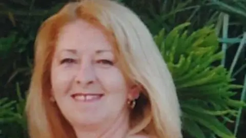 A woman with blond hair, hoop earrings smiles at the camera. There is foliage behind her. It is a close up photo of just her face.