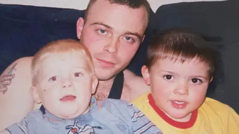 supplied A young man with shaved hair and wearing a black vest cuddles up to two young boys