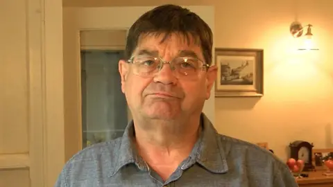 Head-and-shoulders shot of Dr Wilkinson wo has dark hair and silver rimless spectacles on his face. He is wearing a grey shirt and is standing in the hallway of a house