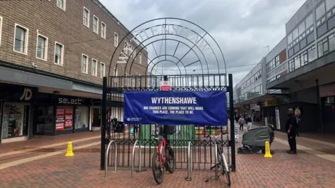 BBC Wythenshawe's current shopping centre, made up of brown brick 1970s-style buildings. Shops including JD Sports and Select are under a dark canopy. A decorative black gate in the middle has a blue canvas sign on it that reads Wythenshawe: Big changes are coming that will make this place the place to be. Two bikes are locked up in front of the gate. 