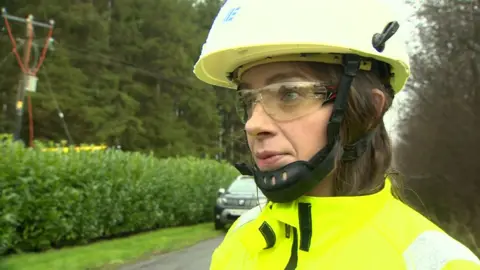 NIE engineer Caron Malone wearing a yellow high vis jacket, safety goggles and a white hard hat. There is a road and hedge behind her. Behind the hedge are trees and power lines. 