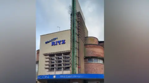 The New Ritz Collective Exterior shot of the Beacon Ritz. It has a round red brick edge and a cream brick frontage. There is a blue logo as well.