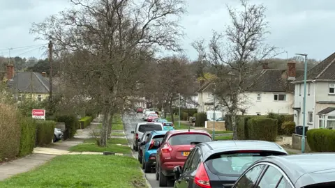 A residential street with white council houses on either side. There are cars parked on either side and trees on the grass verges. It is a grey and cloudy day and there is woodland in the distance.