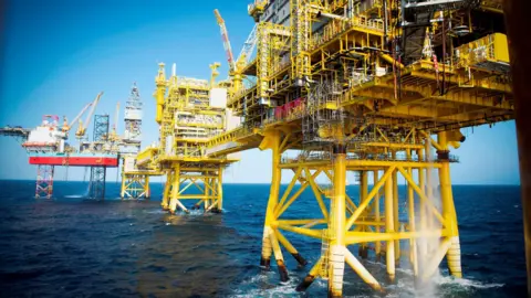 Andy Buchanan/Getty Images Several adjoining production platforms, with yellow legs, sit in the calm North Sea along with a jack-up rig in the distance.