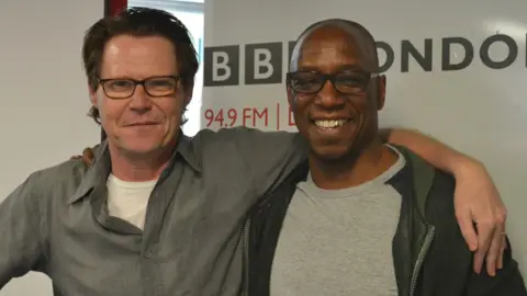 Robert Elms wearing a grey shirt over a white T-shirt, standing next to Ian Wright who is wearing a grey T-shirt an black jacket