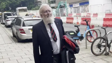 David Wright is pictured outside court. He is wearing a suit and tie and has a long white beard and glasses. He is carrying a bag and coat in his arms. Behind him are bicycles next to a rack and cars parked on the side of a road.