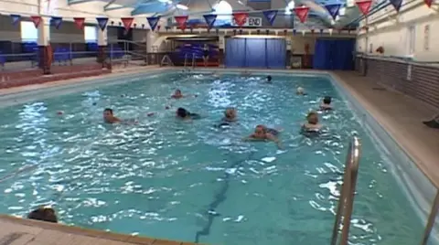 Small indoor swimming pool with some blue and red bunting overhead. There are around a dozen people on the pool