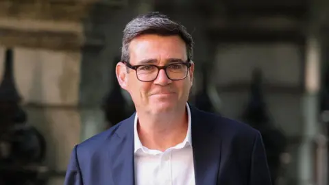 A man with dark hair and black glasses. He is wearing a white shirt and dark blue blazer. 