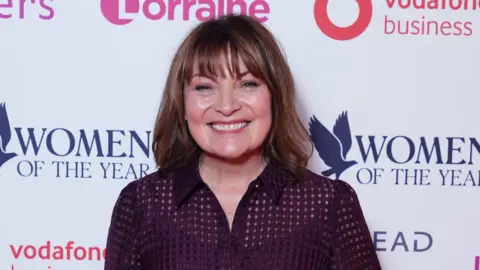 Lorraine Kelly standing in front of an advertising board. She is smiling right at the camera and has shoulder-length dark hair. She is wearing a dark shirt.