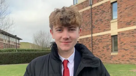 Leo Cavanagh A picture of Leo in college. He has curly light brown hair and is wearing a white shirt and red tie, with a large black coat on. There is a brick building and some grass behind him.