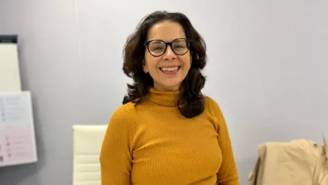 A picture of Education president Andrea Dudley-Owen. She is a women with dark brown mid length hair, wearing a mustard yellow high neck top. she has round glasses on and is smiling.