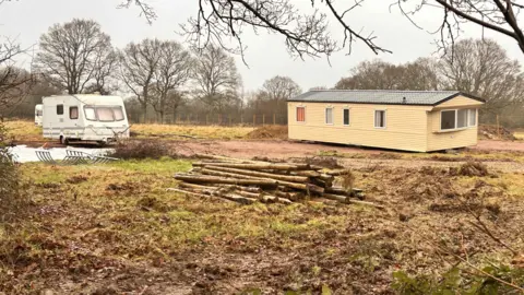 A static and a touring caravan on a field. A large pile of logs lies in the middle of the land.