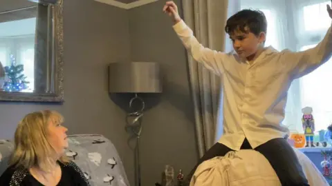 Martin Heath/BBC Boy in white shirt holding his arms up while sitting on pillows in front of a woman who is sitting on a sofa