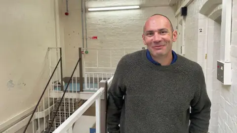 BBC Joel Campbell is standing inside Shrewsbury Prison. He has very short brown hair and is wearing a grey jumper. In the background are some metal stairs.