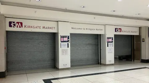 Three closed metal shutters in a shopping centre. Above a sign reads: Kirkgate Market