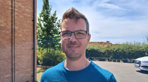 Philip Wood stood in a car park, looking at the camera. He has short brown hair and facial hair and is wearing a blue T-shirt and black-rimmed, rectangular glasses. Behind him in the distance is a large pile of dirt on a construction site.