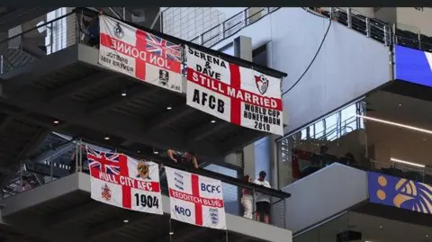 Getty Images A close-up of stands within a football stadium. The picture focuses on two large balconies with St George's flags - red crosses on white backgrounds - draped over the barriers at their edge. The flag at the top right of the image has "still married" printed across the central red stripe. Elsewhere, in the corners, it includes the words "Serena & Dave", "AFCB" and "World Cup honeymoon 2010". The flag also includes the emblem for AFC Bournemouth. 