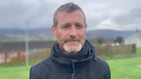 Shay Daly. He has short dark hair and light beard, wearing a black rain jacket. Behind him is grass and fence and other houses.