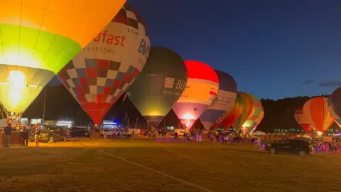 Several colourful hot air balloons lined up with their burners all on at the same time at night. There are colourful lights on the ground, several cars and some people around in the field.