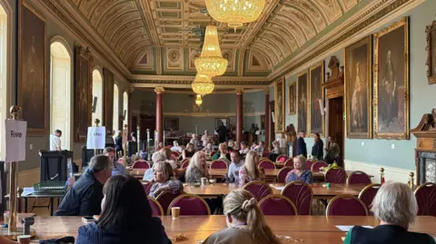Several people sit with their backs to the camera around tables in a large room with paintings on the wall. Empty chairs are visible too and each table has a metal poll with a sign on it to signify which ward it is.