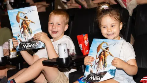 Two children sitting in cinema seats are holding up a Moana film poster each, they both have huge smiles, a can of water each and there's a pack of open Malteasers between them
