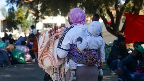 Gallo Images via Getty Images) A woman with a baby strapped to her back is facing away from the camera. She is holding a warm blanket and wearing a mauve headscarf. The baby is wearing a grey hoodie. Some others can be seen out of focus in the background.