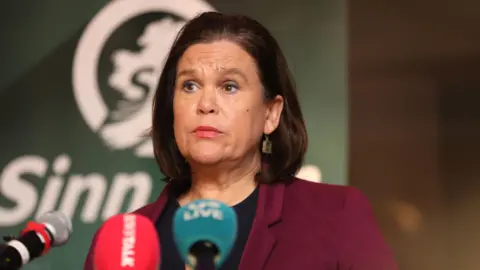 PA Media A woman speaking at a podium with microphones in front of a green Sinn Féin backdrop.