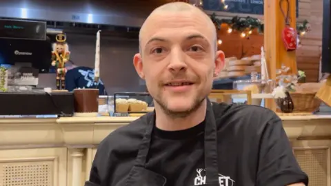 Joe Baugh smiling at the camera. He is sat inside of his cafe and is wearing a black t-shirt and apron.