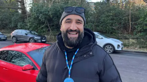 Nathan Turvey/BBC A picture of Sunny Singh wearing a dark coat, hat and and sunglasses on his head, standing next to a red car, with other cars and trees in the background.