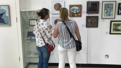 Two women looking at a display of art. 11 pieces of artwork can be seen in the picture, three of them displayed in a glass cabinet, the others on a white wall. 