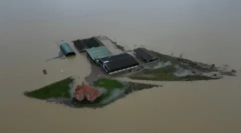 An aerial image of floodwaters surrounding a red brick farmhouse and barns, the water is brown and gives the impression of the farmhouse as an island.