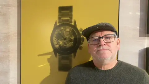 A man wearing a grey jumper and cap. he is stood in front of a yellow canvas bearing a large x-ray image of a watch.