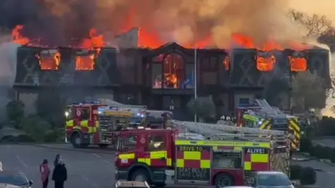 Everything Epping Forest A large building is on fire with its roof collapsed. Clouds of smoke are billowing upwards. Three fire engines are parked in front of the building and a small group of people are watching on.