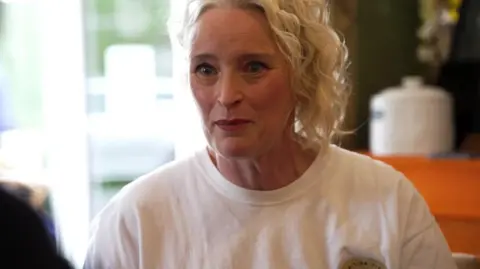 Helen Maxwell, who has a blond hair with a shoulder length and wears white T -shirt, speaks to Roger Johnson, who is outside the camera. She is sitting in a café. 