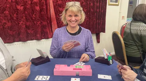 Abbey Smith Abbey Smith sitting at a table playing bridge. A pink plastic box is in front of her, with a blue covering on her table. She is holding cards, with other cards in front of her. There are red patterned drawn curtains. She is wearing a purple top with a black top underneath. She has glasses on her head and has chin-length hair. She is smiling. There are two people at the table with her. A fourth person is sitting down, with their back to the camera, to the right